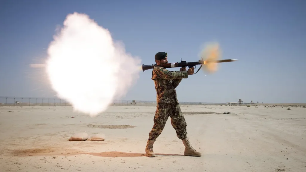 A soldier in camouflage fires a rocket launcher in a desert, producing a huge white backblast cloud.