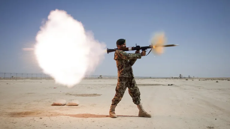 A soldier in camouflage fires a rocket launcher in a desert, producing a huge white backblast cloud.