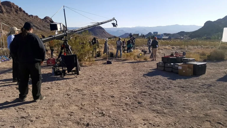 A film crew prepares a large camera crane on a dusty desert mountain location set.