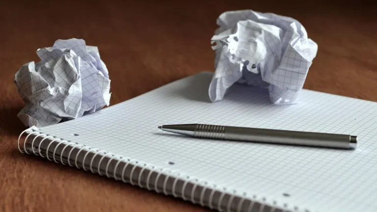 A silver pen rests on a grid-lined spiral notebook beside crumpled paper balls on a wooden desk.