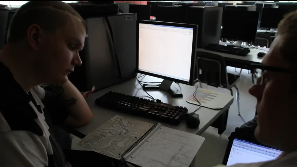 Two young men in a computer lab looking at a notebook with sketches and a computer screen.