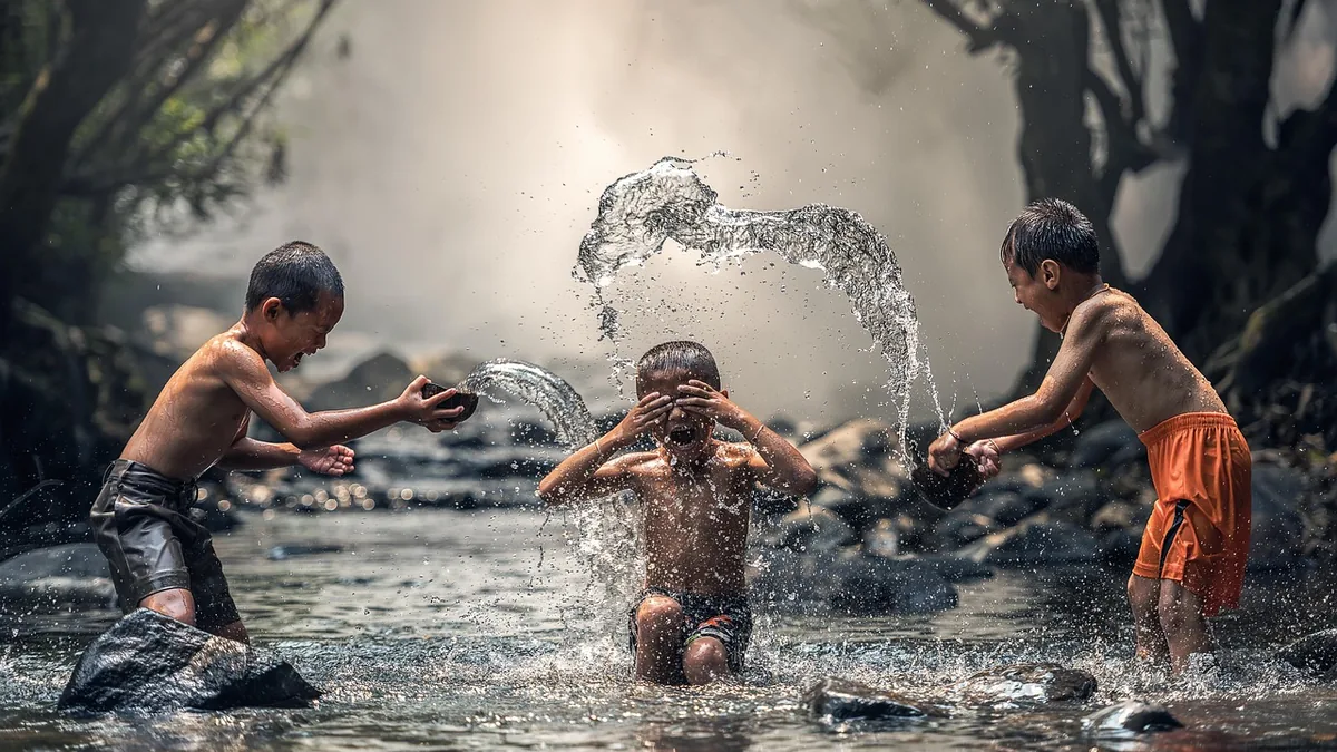 Three children laughing and splashing water on each other while playing in a shallow, rocky river.