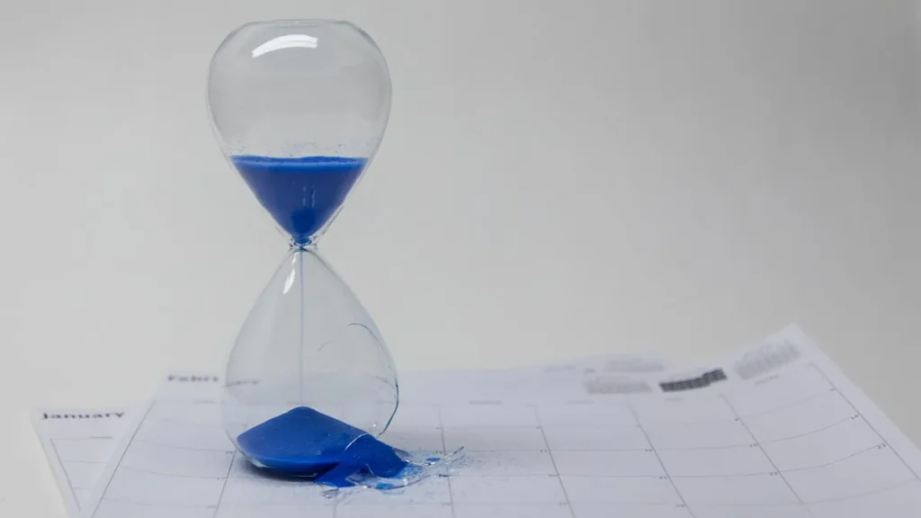 A broken hourglass with blue sand spills onto a calendar, representing a delay in schedule.