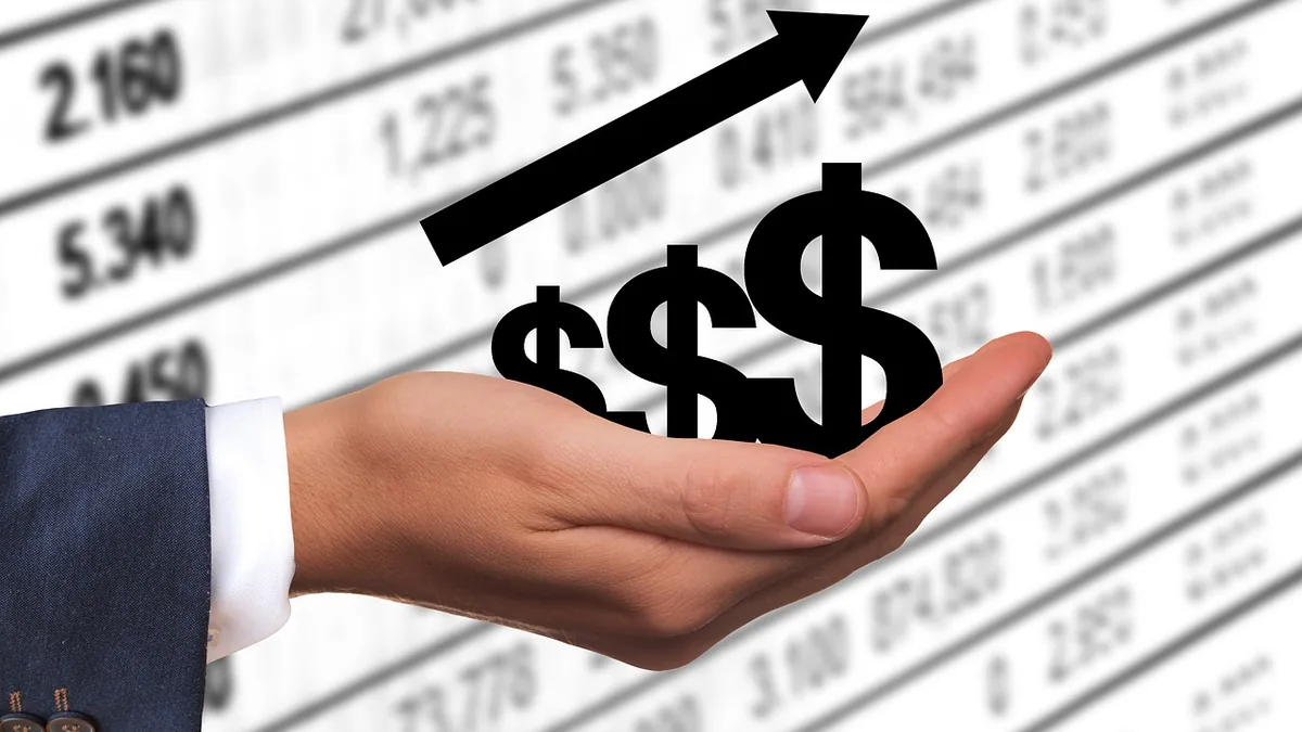 A hand in a suit sleeve holding black dollar signs under a rising arrow over a blurry financial table.