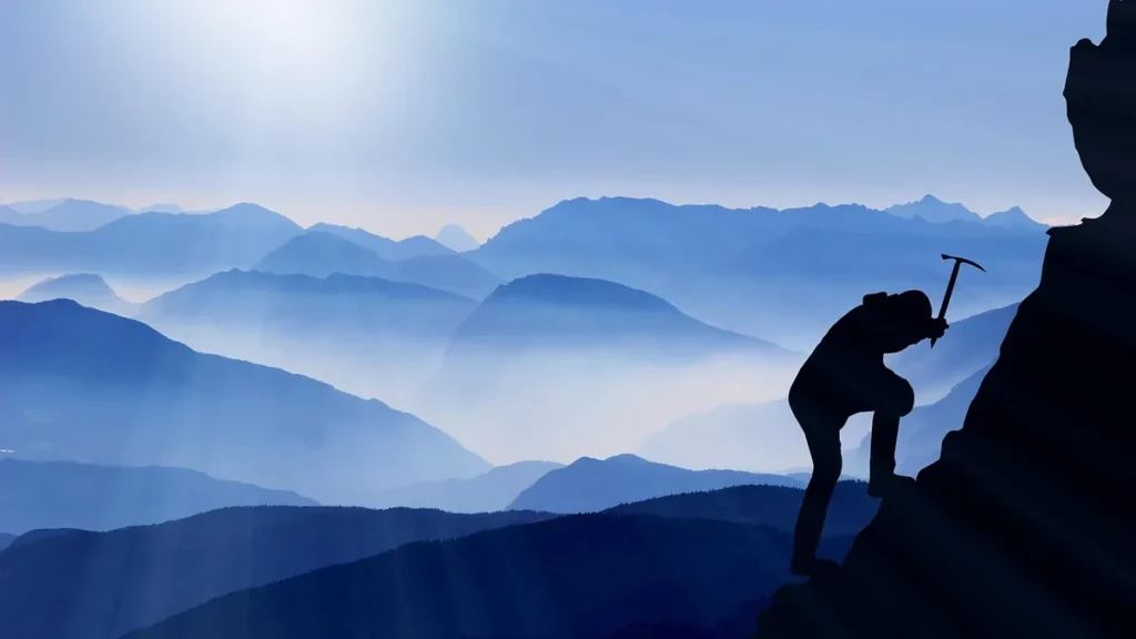 Silhouette of a climber ascending a steep peak against a blue, misty mountain range background.