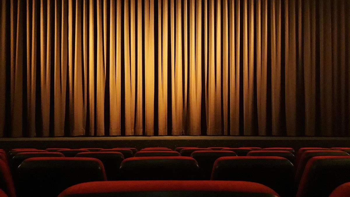 Golden stage curtains closed behind rows of red theater seats under a spotlight.