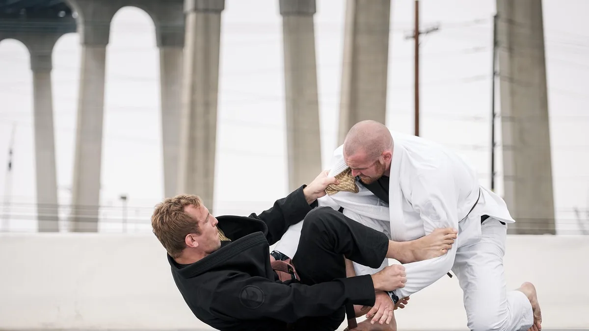 Two martial artists in black and white gis grappling outdoors under giant concrete pillars.