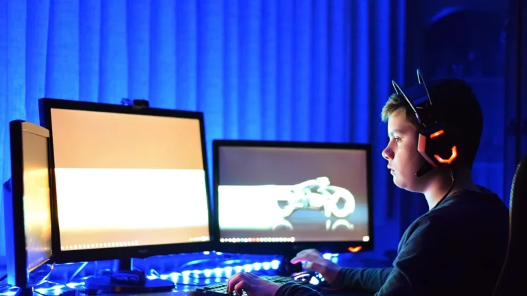 A young gamer sits in profile before multiple monitors under vibrant blue lighting, wearing a glowing orange headset.