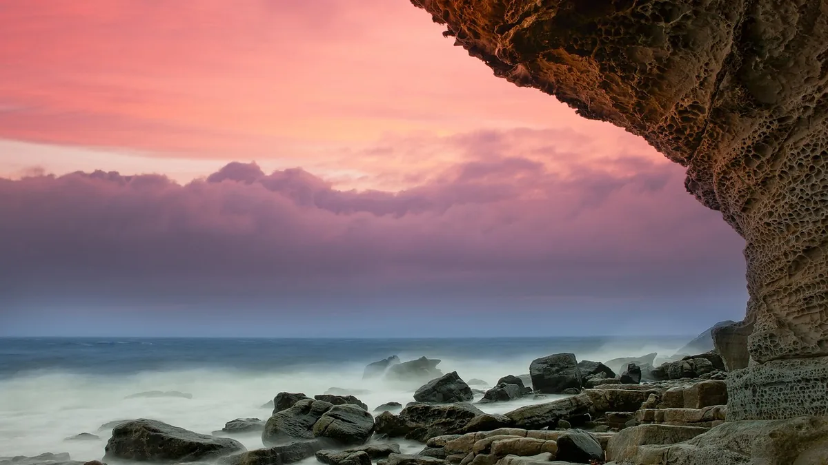 Dramatic pink sunset over a rocky coastline viewed from under a rocky cliff overhang.