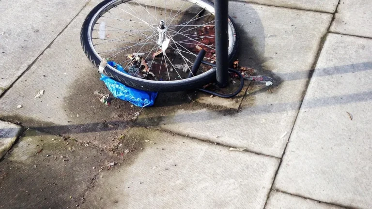 A single bicycle wheel locked to a metal post on a concrete sidewalk with some blue plastic trash underneath.