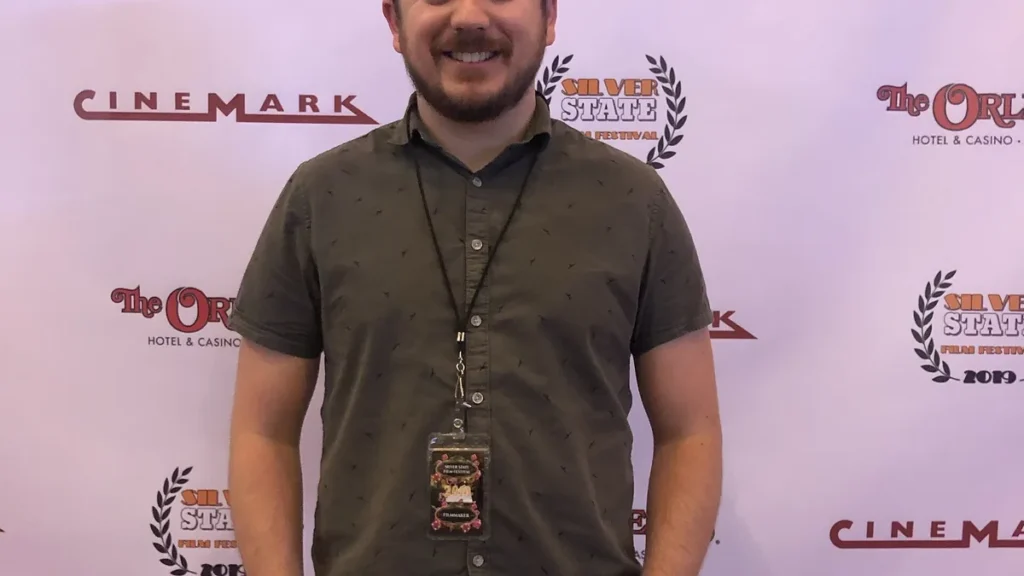 Smiling man in a dark green shirt with a filmmaker badge posing in front of a sponsor backdrop.