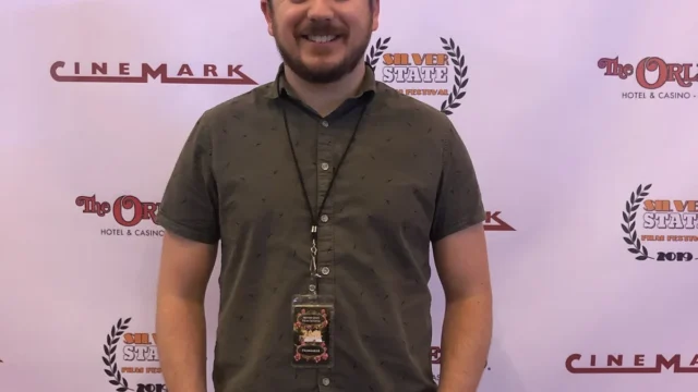 Smiling man in a dark green shirt with a filmmaker badge posing in front of a sponsor backdrop.
