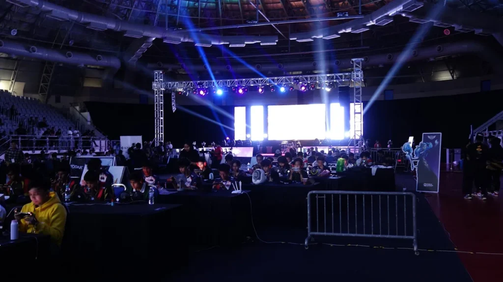 Participants play games at a crowded esports tournament under bright stage lights in a large arena.