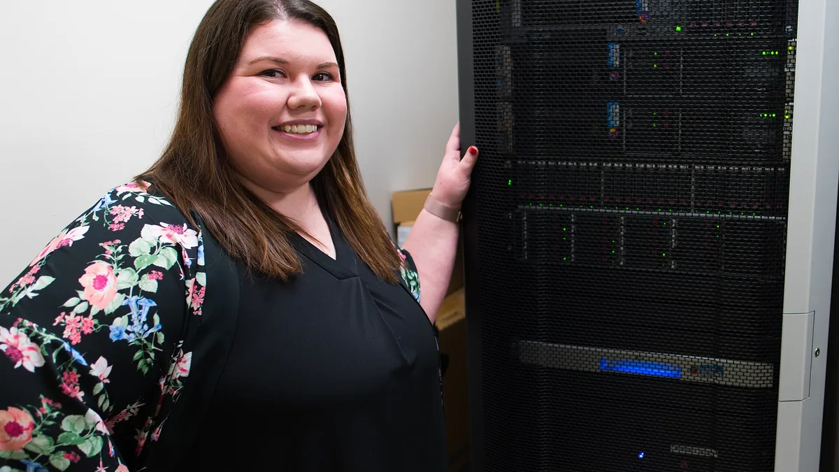 A smiling woman in a black top with floral sleeves stands next to a large server rack with blinking lights.
