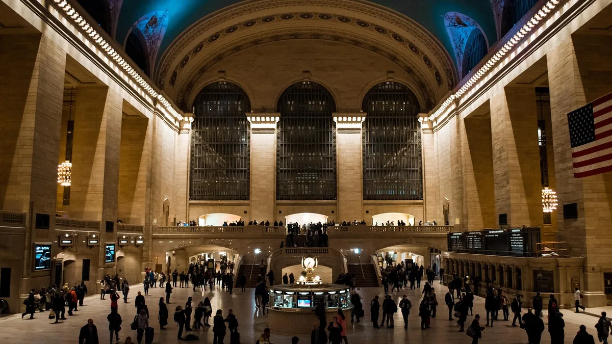 Warmly lit grand interior of Grand Central Terminal with crowds of people walking on the main concourse.