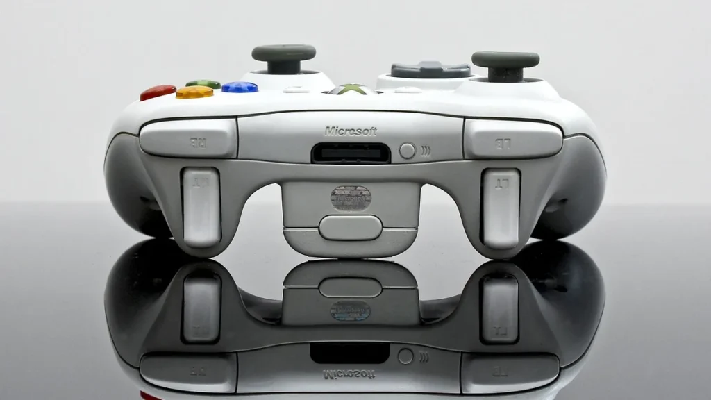 White Xbox 360 controller showing triggers and bumpers, reflected on a glossy black surface.