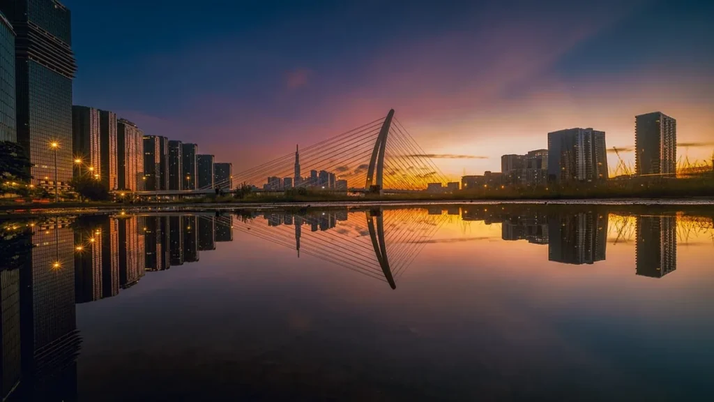 City skyline at sunset with a cable-stayed bridge reflecting in a calm, colorful body of water.