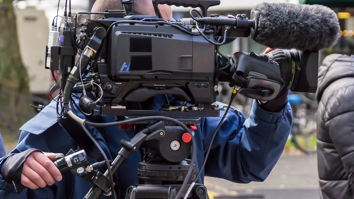 A close-up of a professional broadcast video camera being operated by a crew member in a blue jacket.