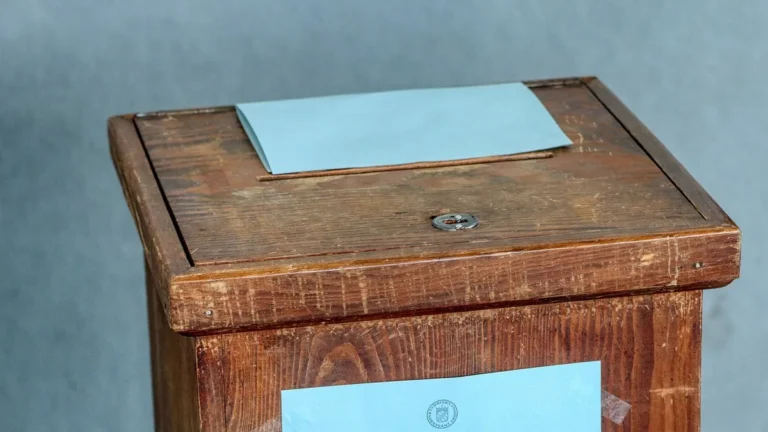 A close-up of a wooden ballot box with a light blue envelope being cast into the voting slot.