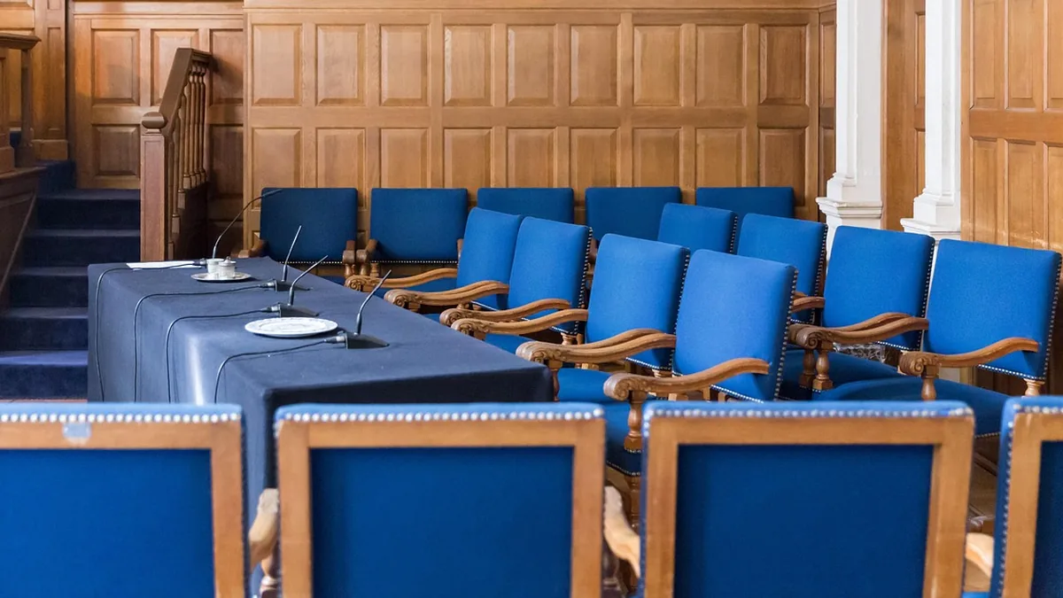 Rows of blue chairs in a wood-paneled tribunal room with a microphone table.
