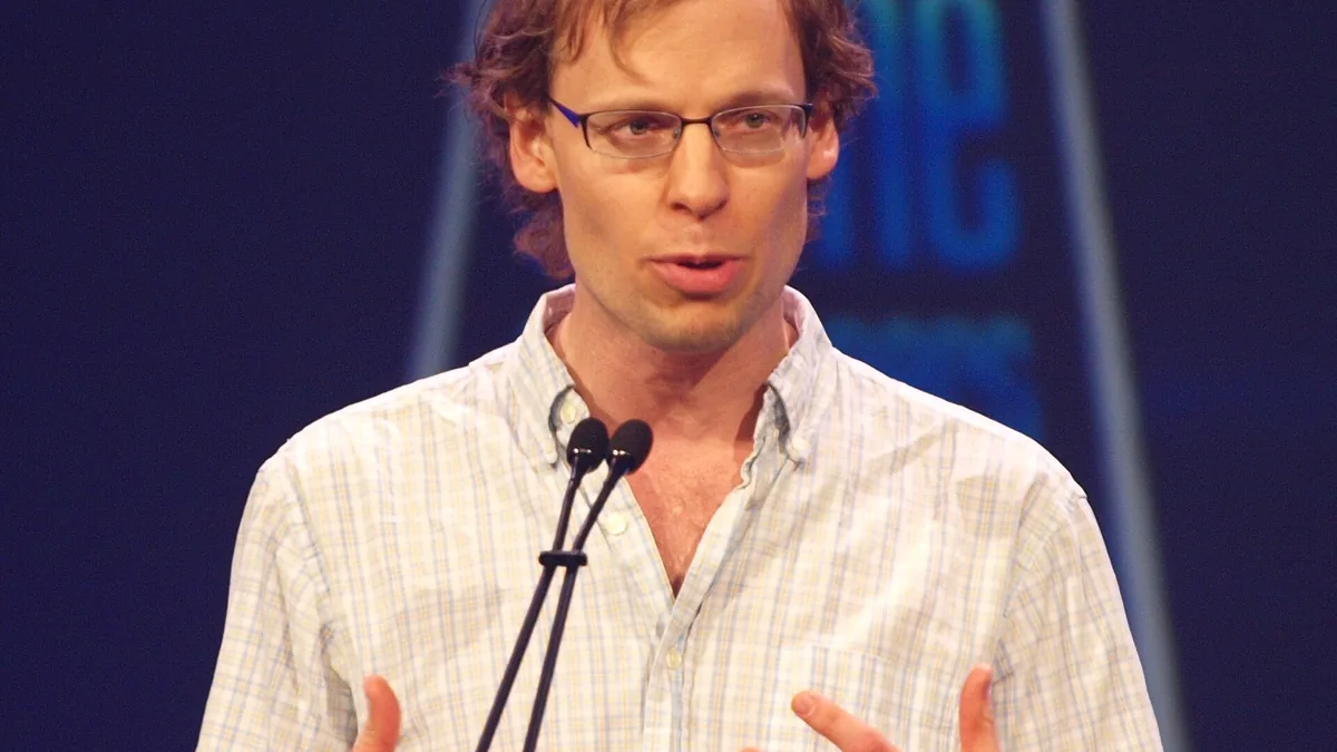 A man wearing glasses and a plaid shirt speaks into dual microphones on a stage.