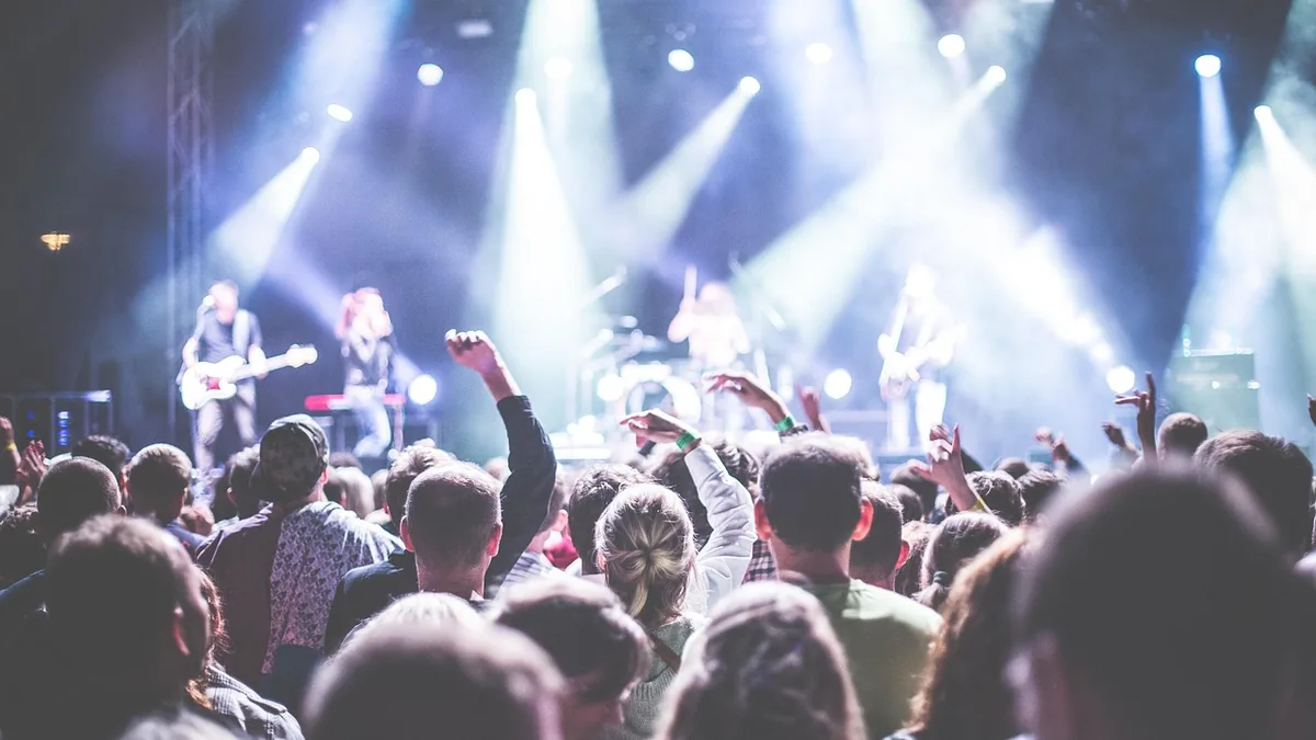 A crowd of people at a brightly lit concert with hands raised, looking towards the stage.