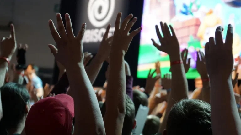 Crowd raising hands at a gaming event, with a colorful screen and a blurry Ubisoft logo in the background.