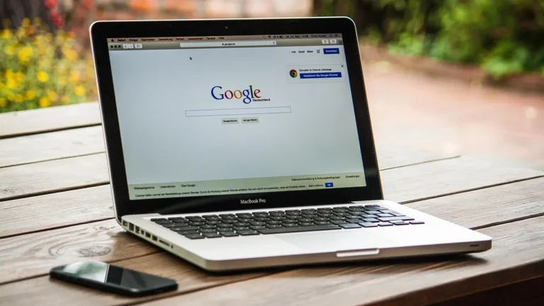MacBook Pro laptop displaying Google search page on a wooden outdoor table with a smartphone nearby.