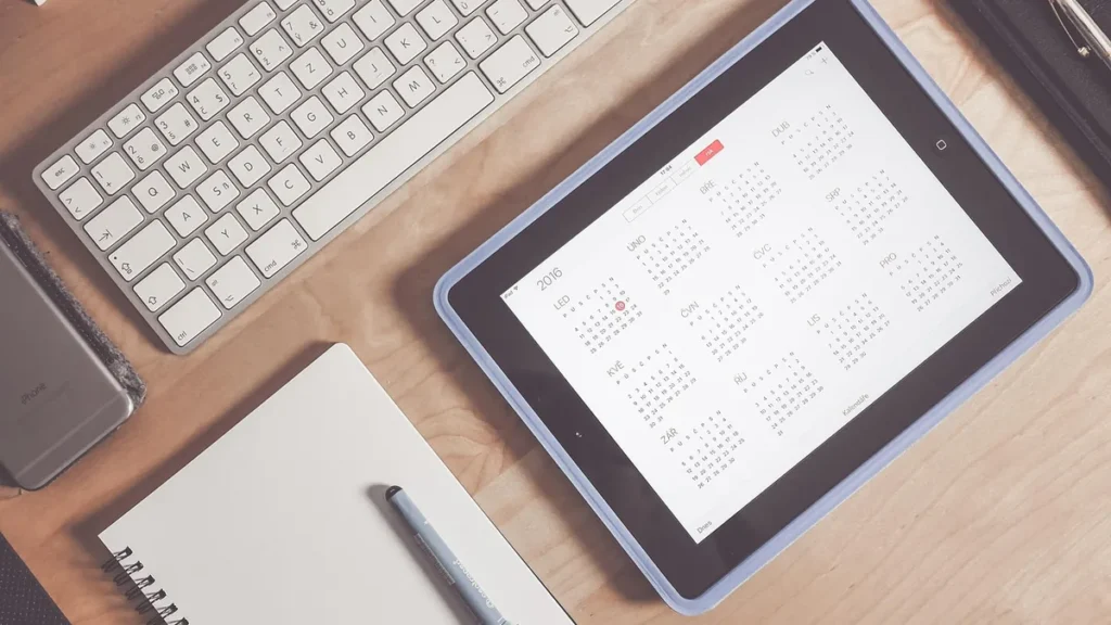 A tablet displaying a 2016 calendar grid next to a keyboard, notebook, and phone on a wooden desk.