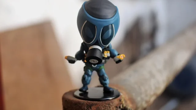 A small blue chibi figurine in a tactical gas mask stands on a rusty pipe.