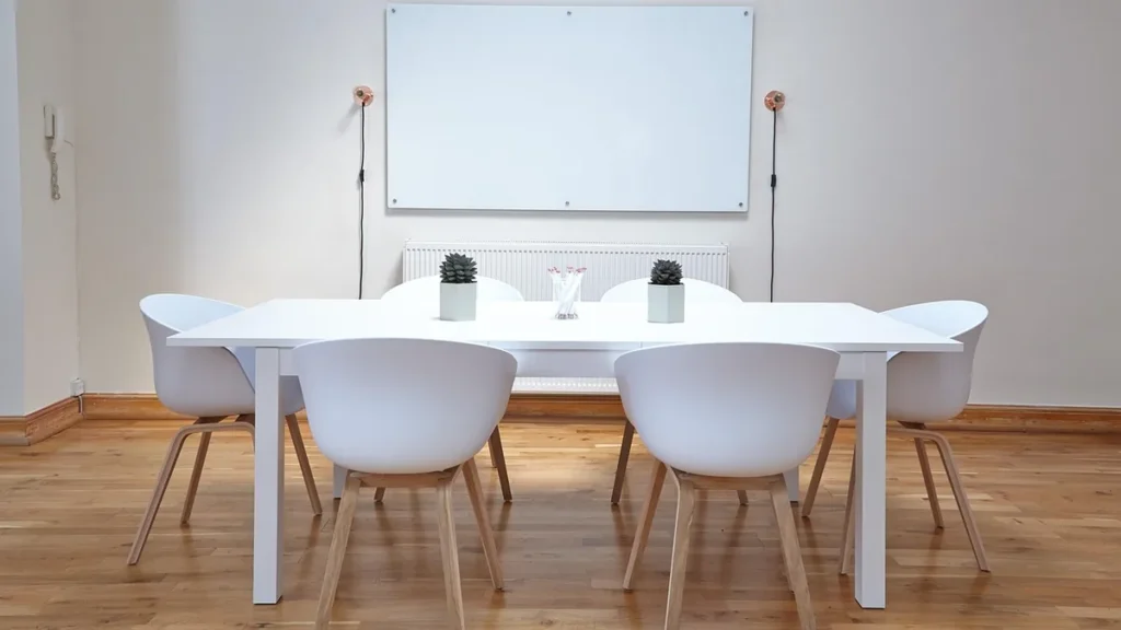 A white conference table with six white chairs in a minimalist office room with a whiteboard on the wall.