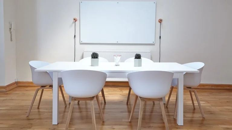 A white conference table with six white chairs in a minimalist office room with a whiteboard on the wall.