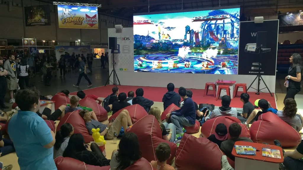 Crowd sitting on red beanbags watching a giant screen play a vibrant fighting game at a gaming event.