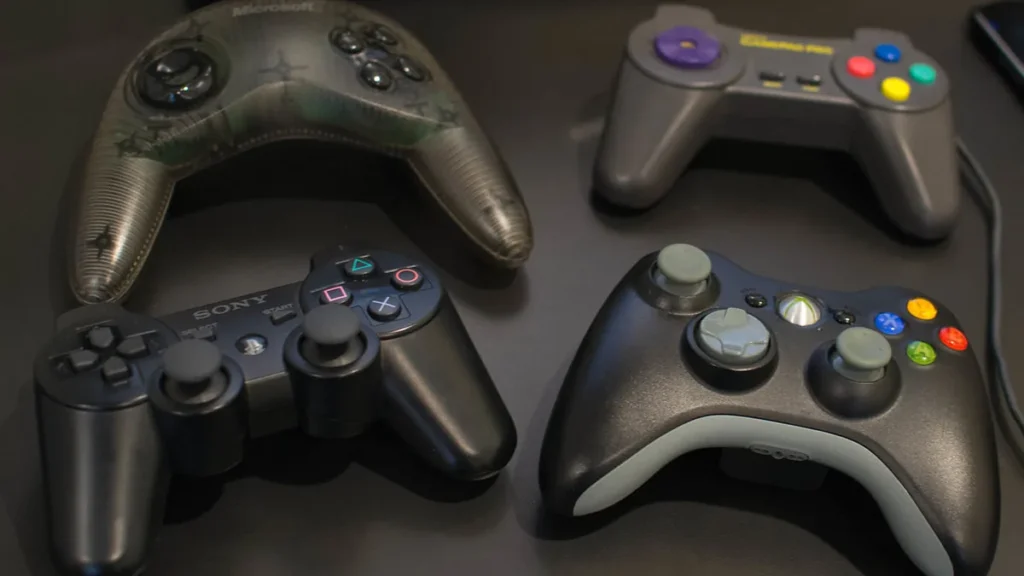 Four video game controllers, including Sony PlayStation and Xbox, on a dark surface.