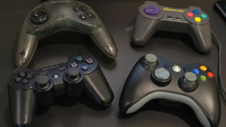 Four video game controllers, including Sony PlayStation and Xbox, on a dark surface.