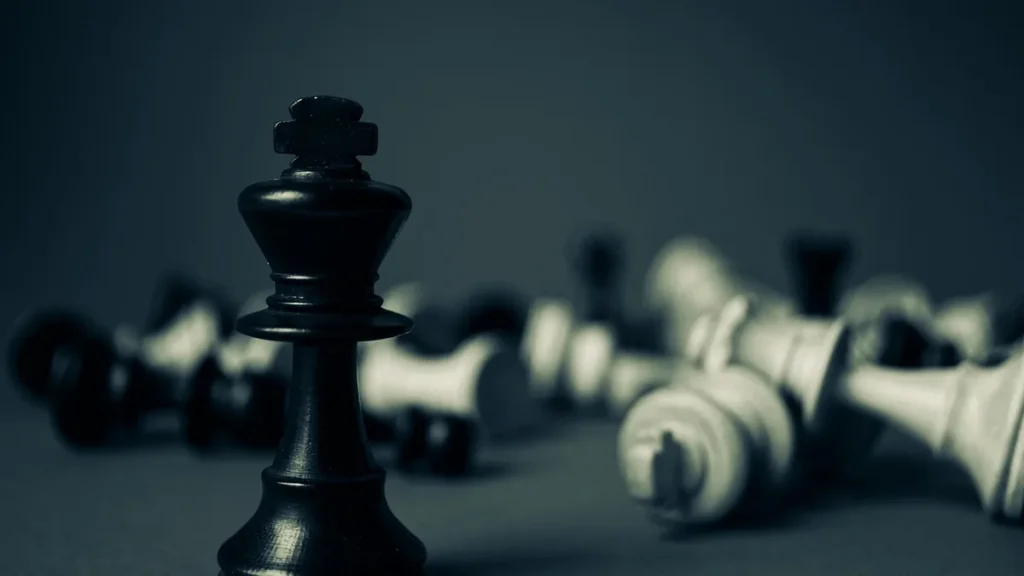 A black chess king stands tall amidst fallen white and black chess pieces on a dark surface.