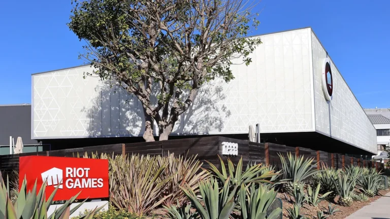 Riot Games headquarters building with a red logo sign, green plants, and a clear blue sky.