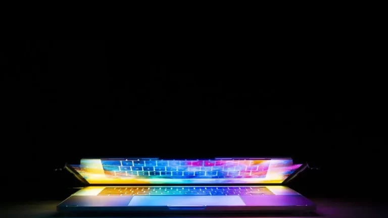 A colorful glowing laptop keyboard and screen reflecting vibrant rainbow colors against a dark background.