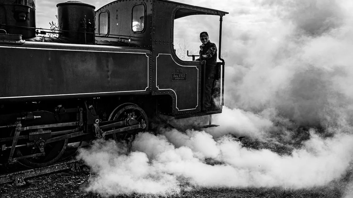 Black and white photo of a vintage steam train driver standing in the cab amidst thick clouds of billowing steam.