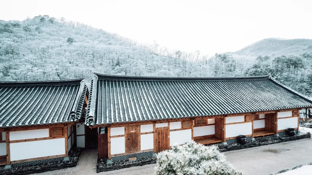 Snow-covered traditional Korean hanok houses nestled against a wintry forest hillside.