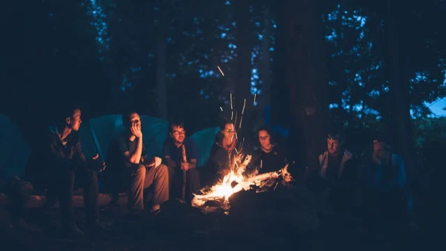 Group of friends sitting around a glowing campfire in a dark forest at night.