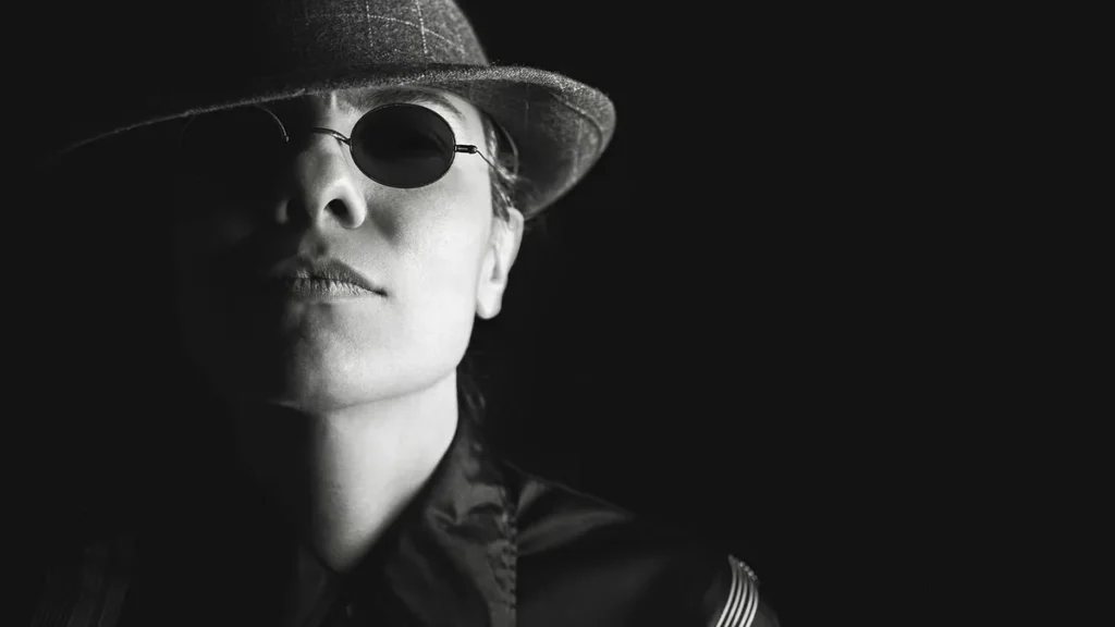 Black and white portrait of a person in a fedora and round sunglasses, cast in dramatic shadow.