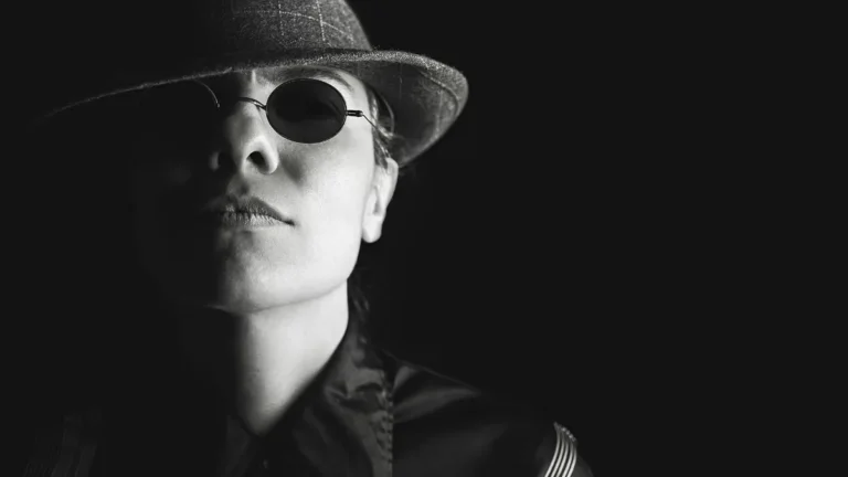 Black and white portrait of a person in a fedora and round sunglasses, cast in dramatic shadow.