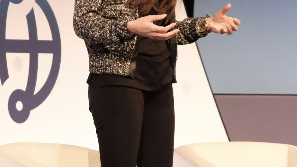 Cropped view of a corporate speaker in a black-and-white jacket gesturing while presenting on stage.