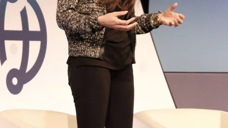 Cropped view of a corporate speaker in a black-and-white jacket gesturing while presenting on stage.