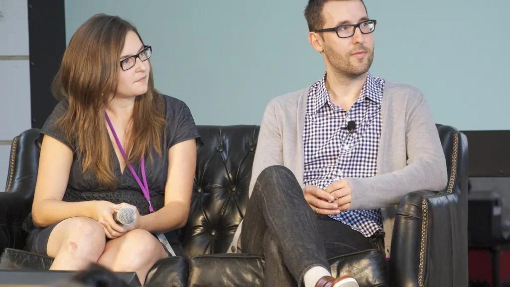A woman and a man wearing glasses sit on a black leather sofa during a panel discussion.