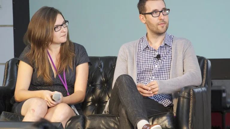 A woman and a man wearing glasses sit on a black leather sofa during a panel discussion.