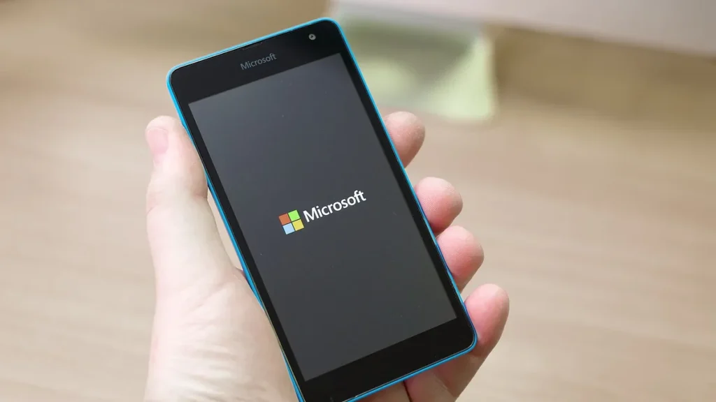 A hand holding a blue Microsoft smartphone displaying the colorful Microsoft logo on a dark screen.