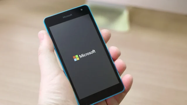 A hand holding a blue Microsoft smartphone displaying the colorful Microsoft logo on a dark screen.