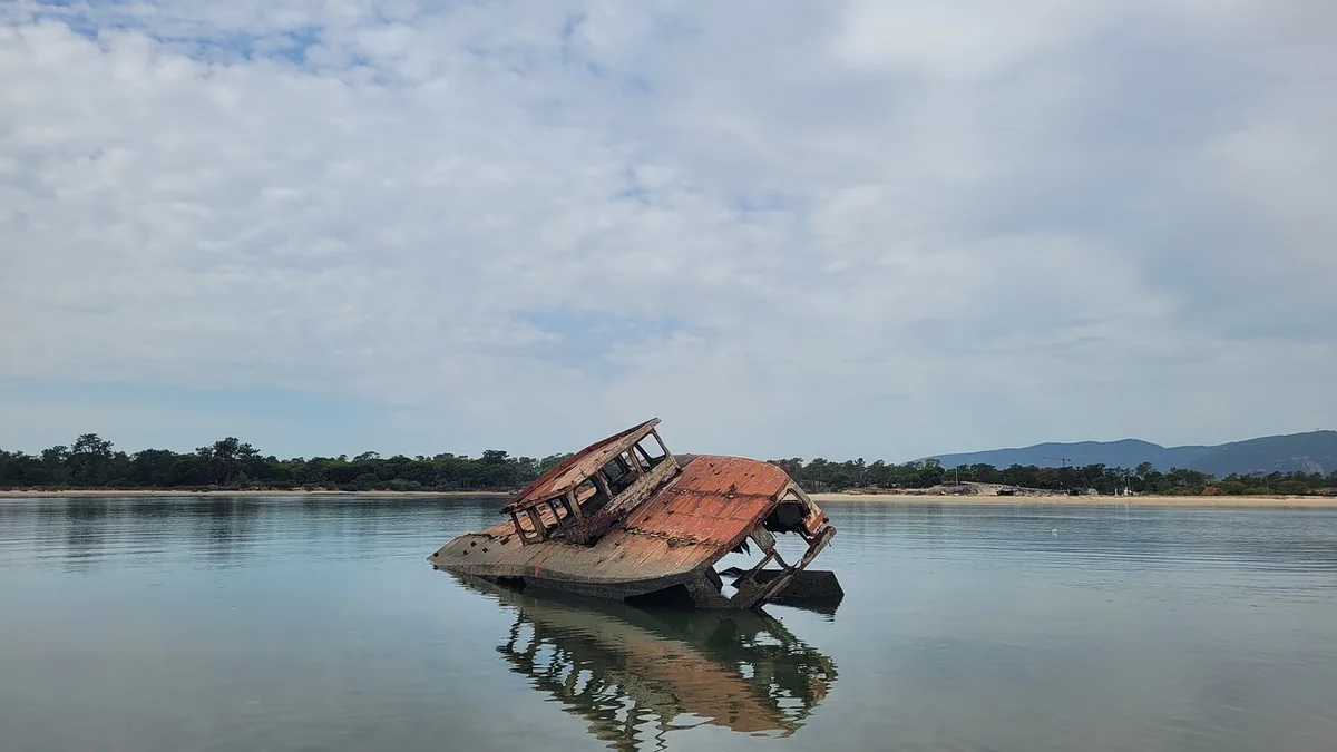 Rusty shipwreck partially submerged in calm water, reflecting cloudy sky and distant shoreline.